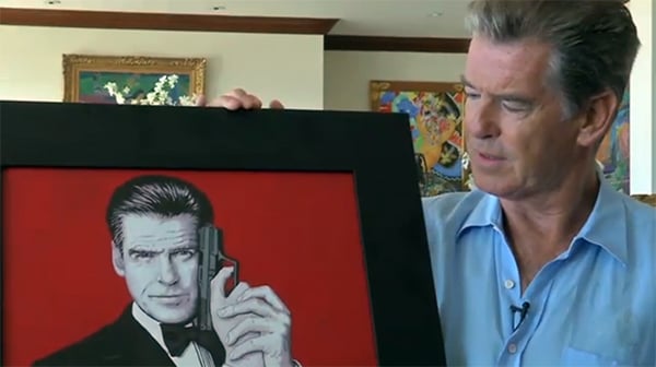 Pierce Brosnan with a portrait of himself that quadriplegic artist Mariam Pare painted using her mouth to hold a paintbrush. Photo: video still from the Mouth and Foot Painting Artists.