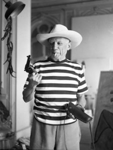 The artist Pablo Picasso, dressed in a striped shirt and sun hat, holds a revolver.