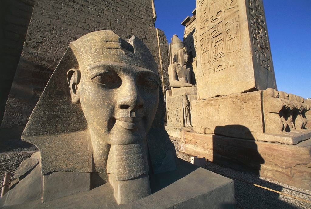 Carved head of Ramesses II with obelisk base, colossal statue of Ramesses II, and first pylon in the background, Temple of Luxor, Thebes (UNESCO World Heritage Site, 1979), Egypt. Egyptian civilization, New Kingdom, 19th dynasty. Photo courtesy of Getty Images.