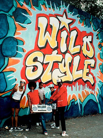 Fab 5 Freddy and Rock Steady Members in front of Wild Style mural by Zypher, Revolt, and Sharp, 1983. Photo by Martha Cooper.