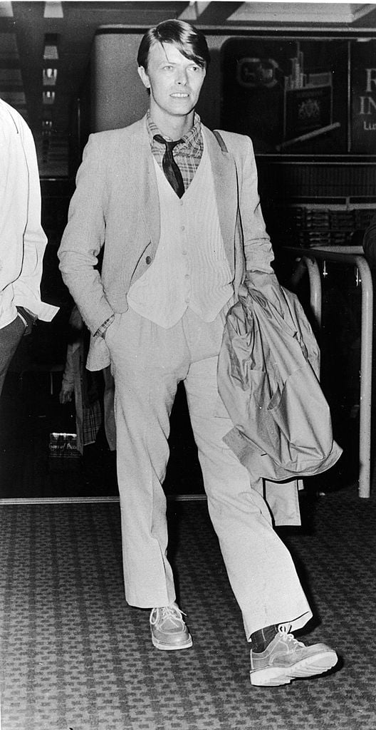 British rock singer and actor David Bowie walks at Heathrow Airport, wearing a suit and sweater and holding his coat, before embarking on a tour of America and Canada, London, England, March 16, 1978. Photo: Express Newspapers/Getty Images. 