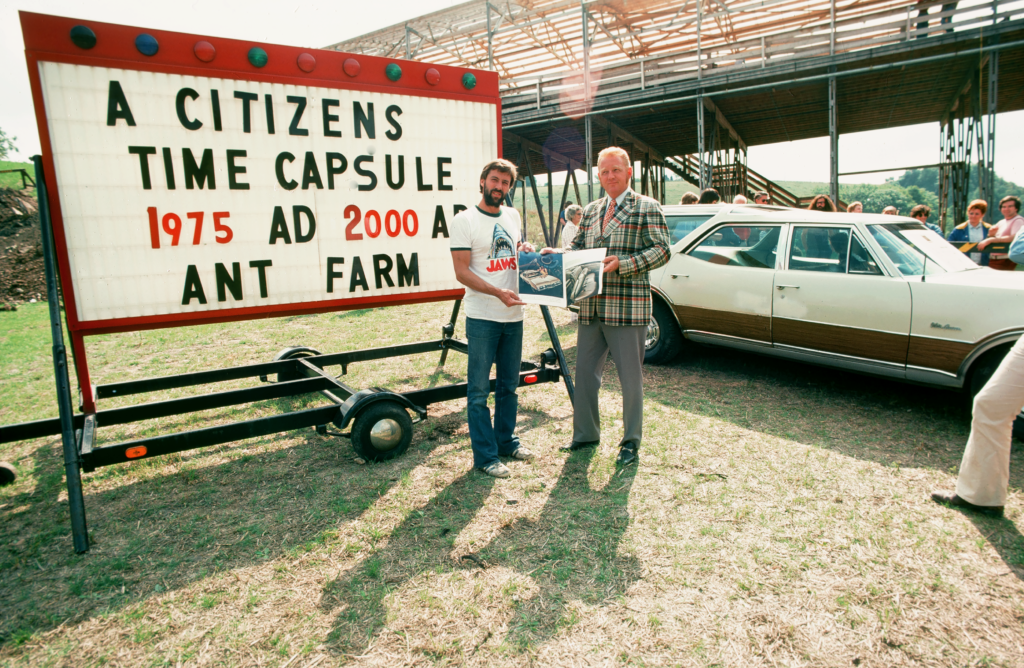 Ant Farm. Citizens Time Capsule, 1975. Doug Michels posing with Oldsmobile Vista Cruiser salesman, holding a 1968 brochure of the Oldsmobile Vista Cruiser station wagon, Lewiston, New York. 35mm slide. Courtesy Chip Lord.