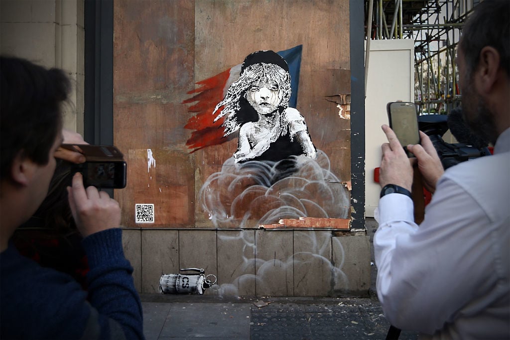 People photograph a Banksy artwork opposite the French embassy on January 25, 2016 in London, England. The graffiti, which depicts a young girl from the musical Les Miserables with tears in her eyes as CS gas moves towards her, criticizes the use of teargas in the 'Jungle' migrant camp in Calais. Photo Carl Court/Getty Images.