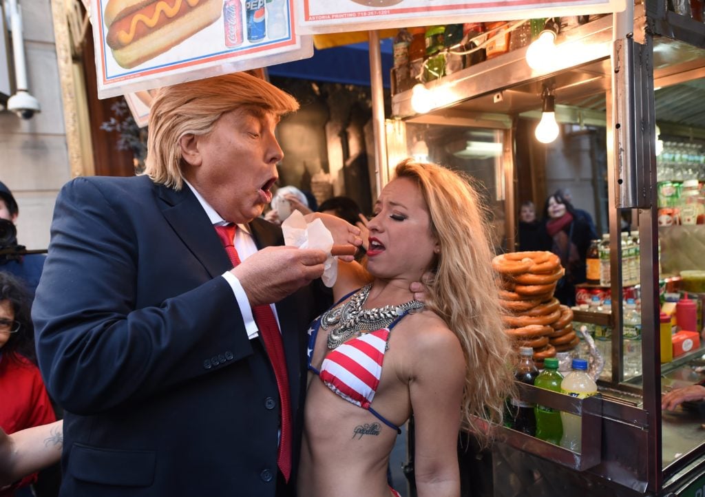 An actor playing Donald Trump as part of an Alison Jackson performance art piece feeds another performer a hot dog. Courtesy of Timothy A. Clary/AFP/Getty Images.