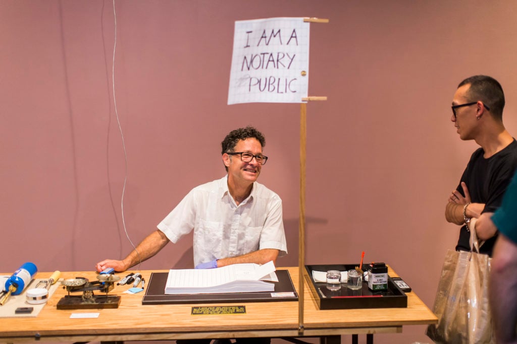 Paul Ramírez Jonas manning the notary table for his piece <em>Alternative Facts</em> in “Paul Ramírez Jonas: Half-Truths” (2017) at the New Museum. Courtesy of the New Museum/Scott Rudd.