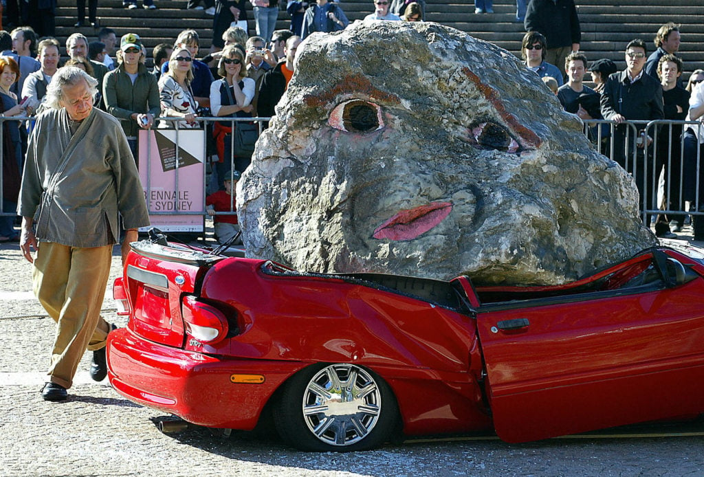American artist Jimmie Durham (L), inspects a car which was crushed with a six tonne granite boulder painted with a human face in front of a large crowd at the Sydney Opera House, 05 June 2004, as a work which forms part of the Biennale of Sydney. The Biennale, Australia's premiere international contemporary visual arts event, is being held between 04 June and 15 August and will feature the work of fifty-one artists from thirty-two countries, with exhibits being shown around Sydney's foreshore, art galleries and museums. AFP PHOTO/Greg WOOD (Photo credit should read GREG WOOD/AFP/Getty Images)
