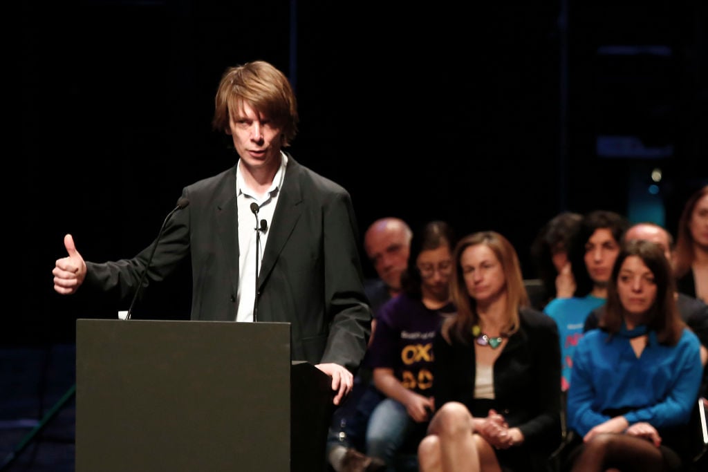 A man behind a lectern speaking into a microphone with a crowd behind him