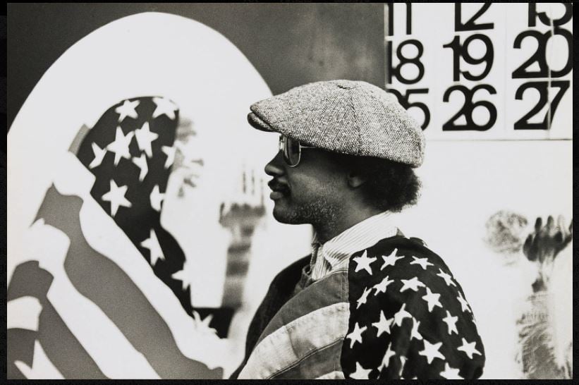 David Hammons in his studio (1970), with <i>Pray for America</i> (1969) shown at left and a portion of <i>Wine Leading the Wine</i> (c. 1969). Photo: Robert A. Nakamura, courtesy of the Hammer Museum.