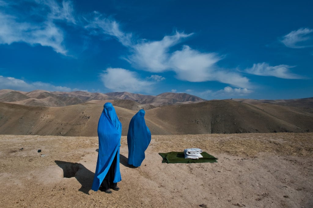 Lynsey Addario, Two women on side of road, from Weha village, four hours in car to clinic. The father has lost two wives already, and has taken a third wife, half his age. His name is Shir Mohammad, and his wife, in burqua on hill, is Noor Nisa, 20. it is her first pregnancy, and her water has just broken and their car broke down on the side of the road. Shir mohammad went to look for other transport, and Noor Nisa and her mother, Nazer Begam, 40, are waiting transport to the hospital. Courtesy of the International Center of Photography.