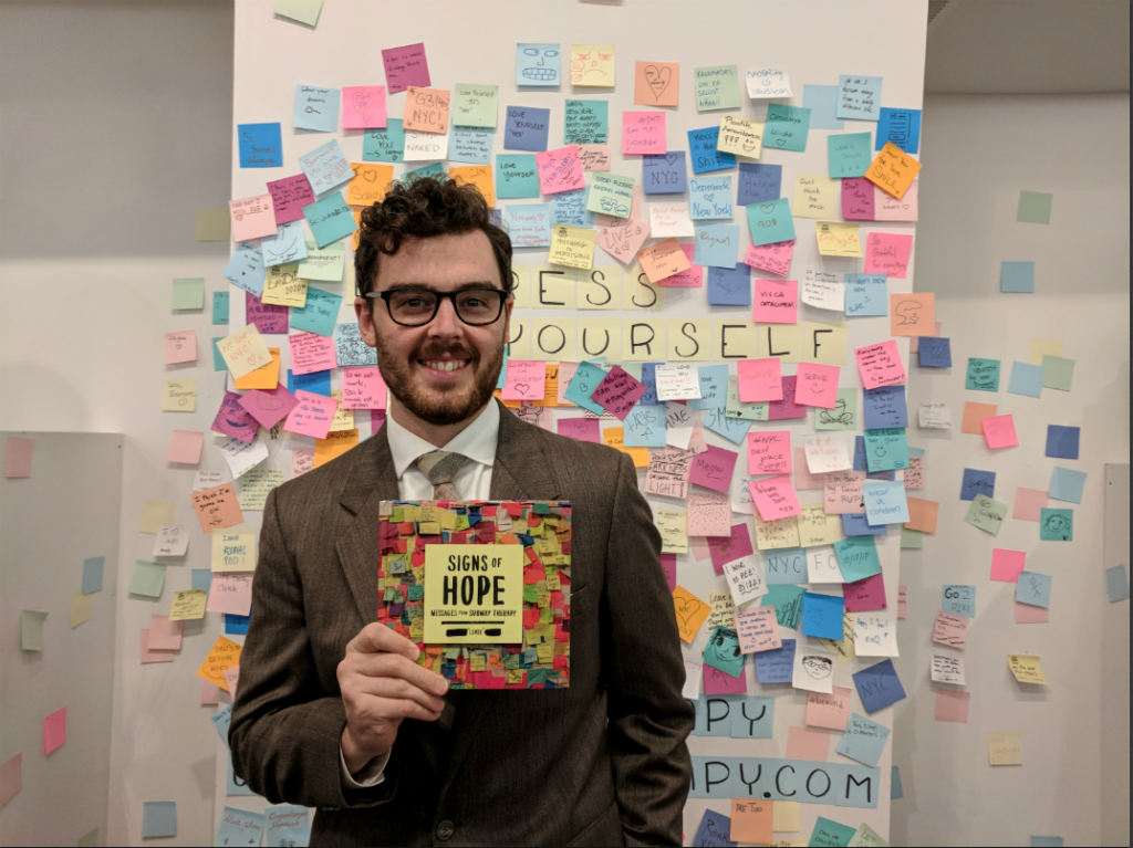 Matthew "Levee" Chavez poses with his book <i>Signs of Hope</i> in front of his installation at Artists & Fleas, (2017). Courtesy of Hannah Pikaart.