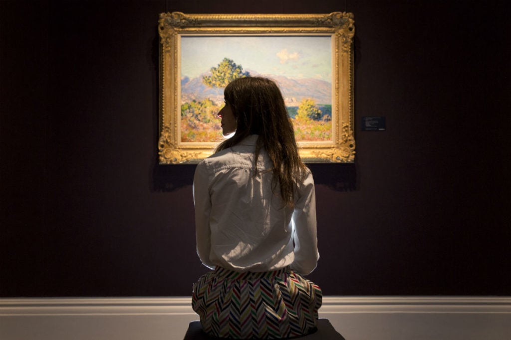A woman sits in front of Claude Monet's 1888 piece <em>Antibes, vue du plateau Notre-Dome</em>,estimated to sell for £6-£8 million, on display at Sotheby's auction house on June 18, 2014 in London, England. Photo by Rob Stothard/Getty Images.