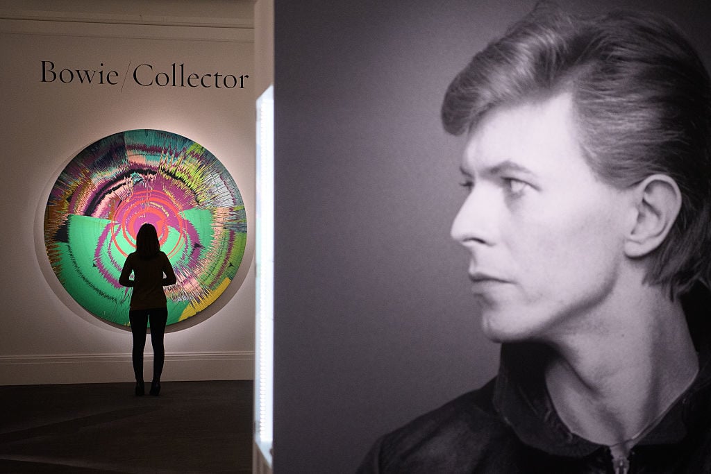 A gallery assistant poses with <em>Beautiful, Hallo, Space-Boy Painting</em> by Damien Hirst with David Bowie (est. £250,000-350,000) during the press preview of the "Bowie/Collector" auction at Sotheby's on November 1, 2016 in London, England. Photo by Leon Neal/Getty Images.