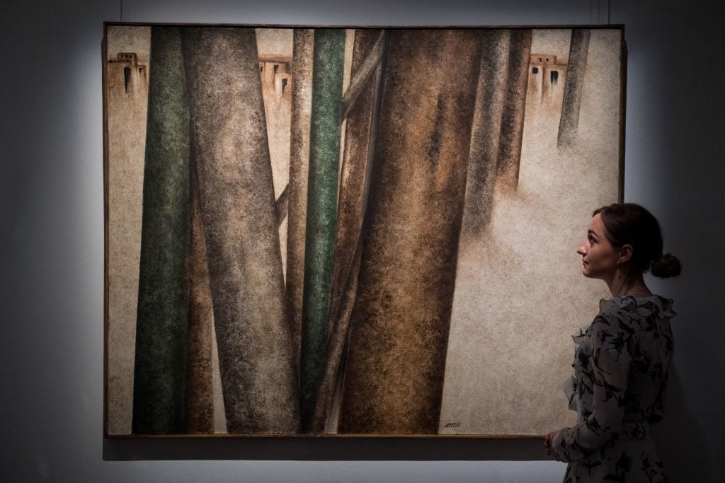 Sotheby's employee poses next to <em>Untitled (Tree trunks and village scene)</em> by Sohrab Sepehri during a press preview of Orientalist and Middle Eastern Art Week at Sotheby's on April 20, 2018 in London, England. Photo by Chris J Ratcliffe/Getty Images for Sotheby's.