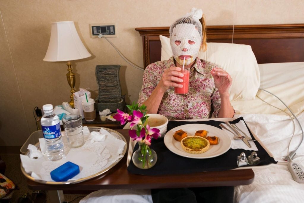 Dr. Steven Teitelbaum’s forty-nine- year-old patient, who is recovering from a full-face laser resurfacing and an upper eye lift, eats lunch in a private room at Serenity, a luxury aftercare facility that offers private chefs, spa treatments, and twenty- four-hour nursing, Santa Monica, California, 2006. Featured in Lauren Greenfield's <em>Generation Wealth</em>. Photo courtesy of Amazon Studios ©Lauren Greenfield, all rights reserved.