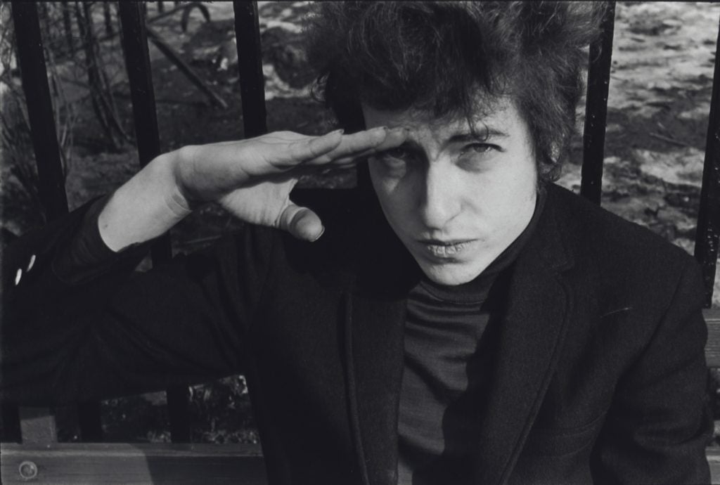 Fred W. McDarrah, Bob Dylan, sitting on a bench in Christopher Park (across the street from the offices of the Village Voice since 1960), either salutes or shields his eyes from the sun, January 22, 1965. Photo courtesy of Steven Kasher Gallery.