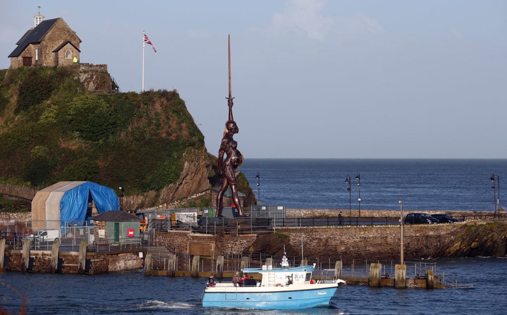A boat passes in front of Damien Hirst's bronze sculpture of a pregnant woman in Ilfracombe, England. The bronze-clad, sword-wielding 65-foot statue, named <em>Verity</em>, has been controversially given to the seaside town by the artist on a 20-year loan. Photo by Matt Cardy/Getty Images.
