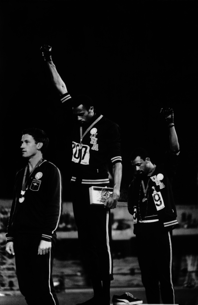 A photo of Tommie Smith fellow medalist and John Carlos looking down and raising his fist as he stands on the podium at the 1968 Olympics. Photo ©Time & Life Pictures/Getty Images.