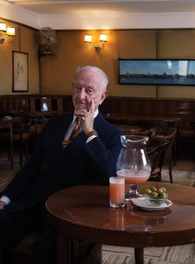 Arrigo Cipriani at Harry's Bar, Venice. He is an old balding white man in a suit, with a pitcher and glass of the pink peach bellini, sitting in a wood paneled restaurant.