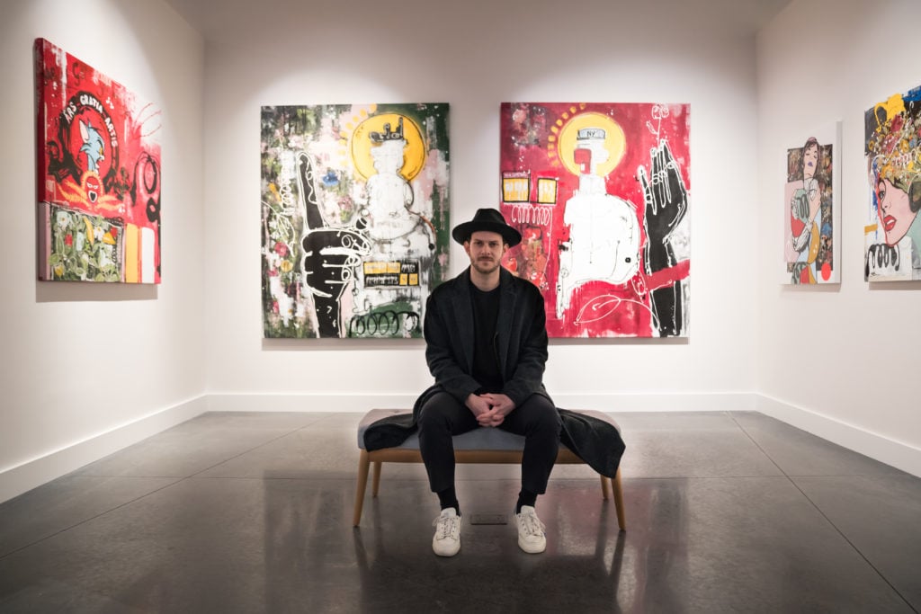 Artist Felipe Echeverry known as LaSso seated in front of his paintings, 2019. Courtesy Avant Gallery. 
