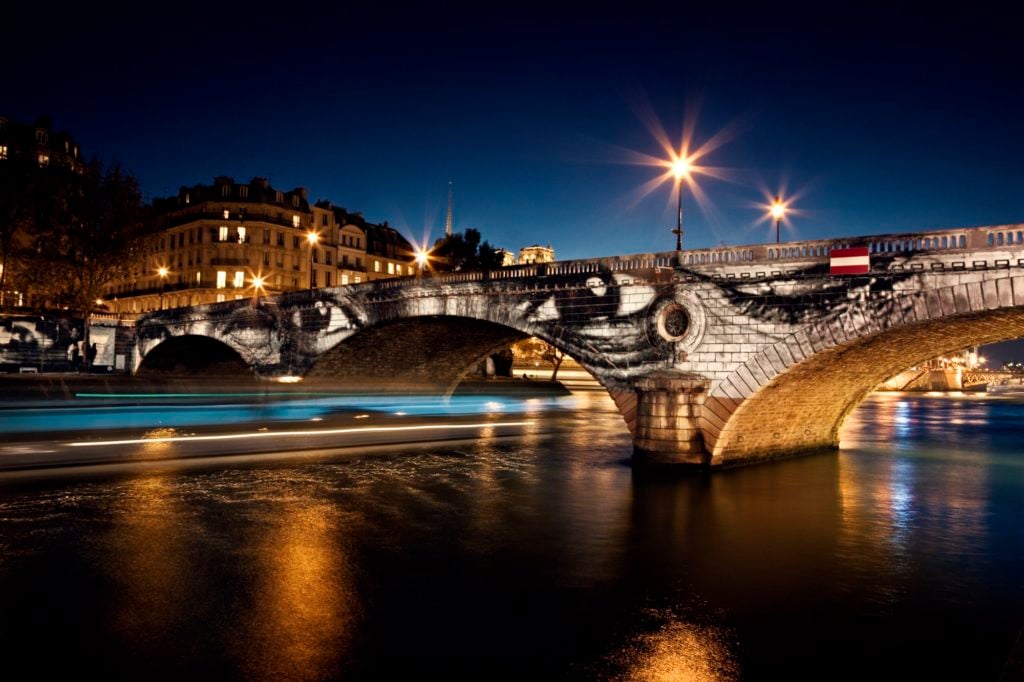 JR, <em>28 Millimètres, Women Are Heroes, Exhibition in Paris, Pont Louis-Philippe–Pont Marie Side by Night, with Barge, France</em> (2009). Photo ©JR- ART.NET.