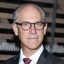A headshot of an older bald white man with thick-rimmed glasses in a suit and tie with a white-collared shirt, photographed in front of a horizontally striped wall from the shoulders up.