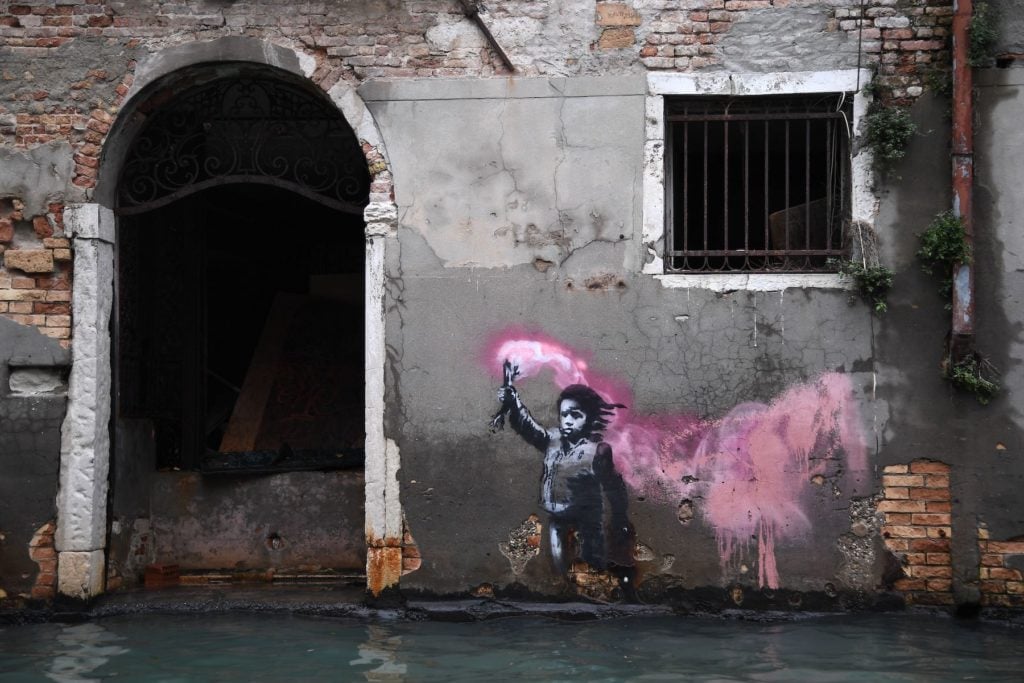 An artwork by street artist Banksy portraying a migrant child wearing a lifejacket and holding a neon pink flare pictured during the November 13, 2019 floods in Venice. Photo by Marco Bertorello/AFP via Getty Images.An artwork by street artist Banksy portraying a migrant child wearing a lifejacket and holding a neon pink flare pictured during the November 13, 2019 floods in Venice. Photo by Marco Bertorello/AFP via Getty Images.
