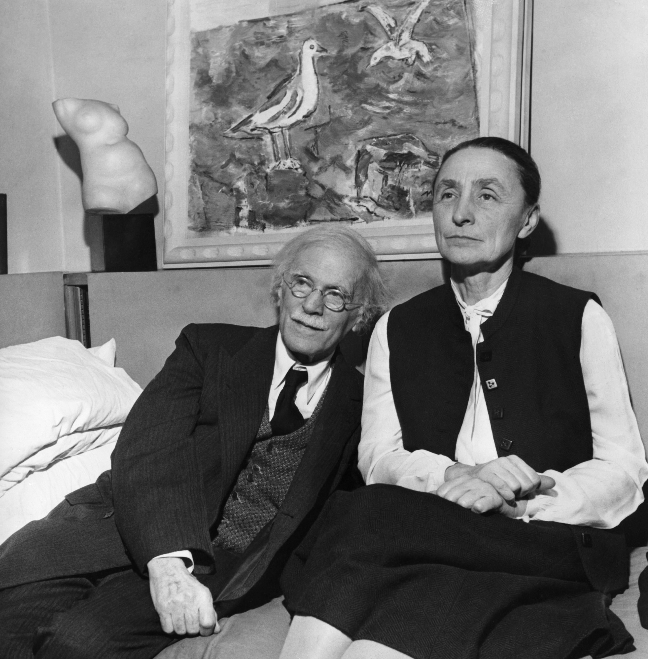 a black and white portrait of the elderly Alfred Steglitz sitting on a sofa next to his wife, Georgia O'Keeffe. A sculpture and a painting are in the background.