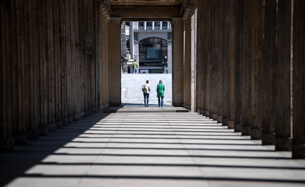Two women walk through a colonnade in front of the Alte Nationalgalerie and the Neues Museum on Museum Island. Photo: Bernd von Jutrczenka/picture alliance via Getty Images.
