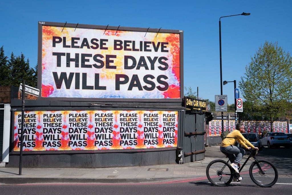 A cyclist rides past a billboard reading 'please believe these days will pass' in Shoreditch, east London. Photo by Dominic Lipinski/PA Images via Getty Images.