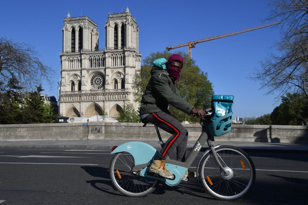 A man wearing a protective mask rides his bicycle in front of Notre Dame Cathedral in Paris after repair work stops due to the global health crisis. Photo by Stephane Cardinale, courtesy of Corbis/Corbis/Getty Images.