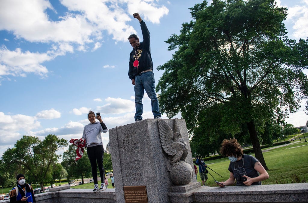 A man stands with his fist raised on the spot where a statue of Christopher Columbus, which was toppled by protesters, stood on the grounds of the State Capitol on June 10, 2020 in St Paul, Minnesota. Photo by Stephen Maturen/Getty Images.