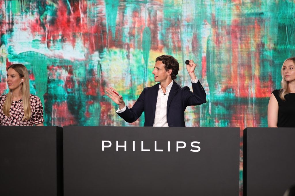 Auctioneer Henry Highley in the Phillips saleroom in London, flanked by real people but talking mostly to images on large screens. Photo courtesy Phillips.