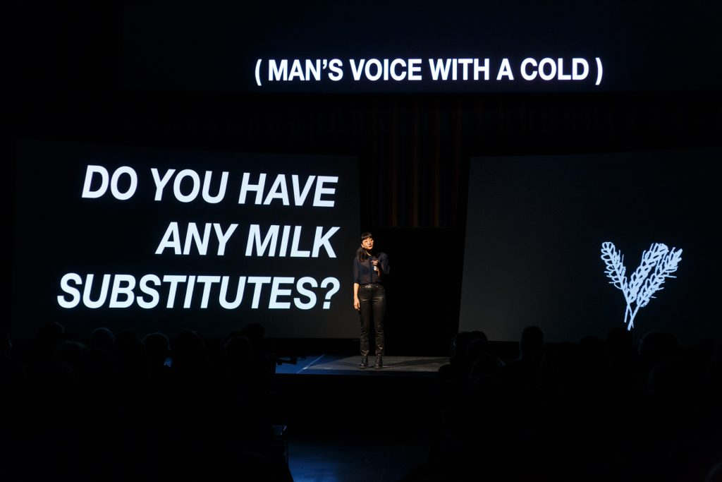 Dressed in a sleek black outfit, Christine Sun Kim stands on stage among three large, black screens. The left screen reads, "DO YOU HAVE ANY MILK SUBSTITUTES?" On the right screen, a drawing of wheat stocks. Above her, a sound description reads, "MAN'S VOICE WITH A COLD."