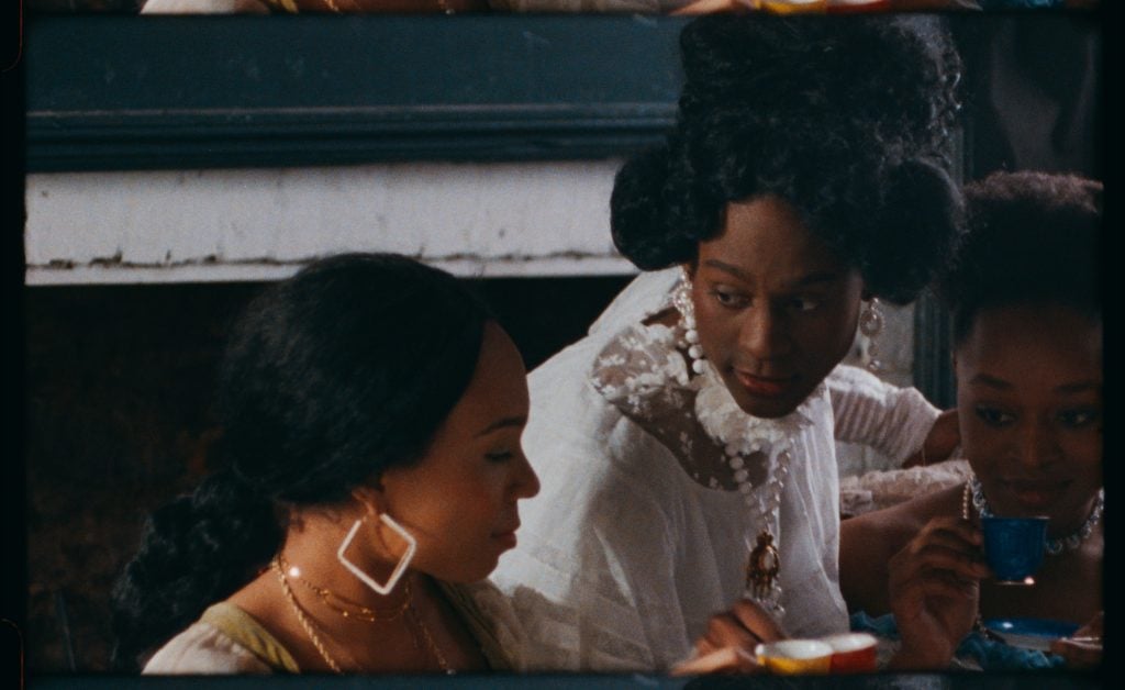 Film still. Rowin Amone plays Mary Jones, wearing a white dress and white pearls with her black hair in a sculptural updo. She leans forward, in conversation with two other lavishly dressed Black women on either side of her.