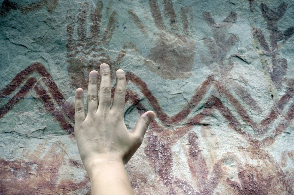 Palaeo-anthropologist Ella Al-Shamahi compares her hand to a handprint created around 12,000 years ago at Cerro Azul in Guaviare state, Columbia. Photo by Marie-Claire Thomas, courtesy Wild Blue Media.