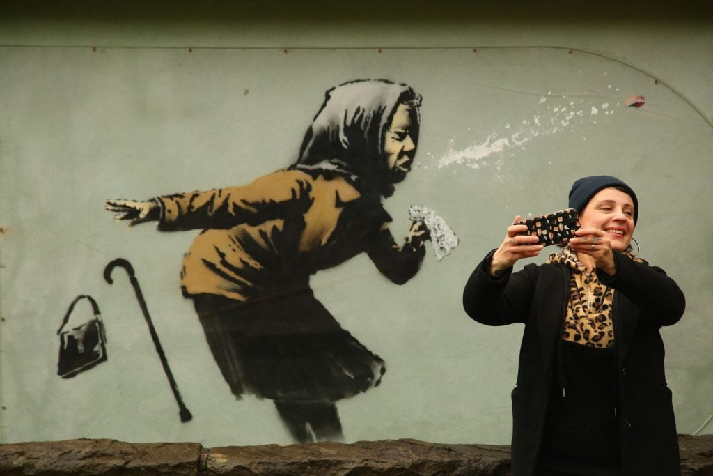 A person photographs a mural created by British artist Banksy entitled "Aachoo!!" showing a woman wearing a headscarf sneezing and dropping their handbag and cane, in Bristol. (Photo by Geoff Caddick/AFP via Getty Images.)