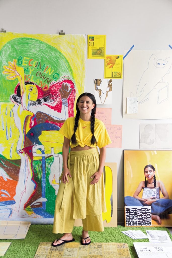 KT Pe Benito in their former live/work space in Queens, New York, standing in front of their oil pastel, <em>Becoming My Own Island</em> (2019), and excerpts from their multimedia installation <em>Entries to Faustina (Growing out of colonialism for my grandmother’s sake</em> 2018. Photo by Sunny Leerasanthanah, ©2021 Jasmin Hernandez.