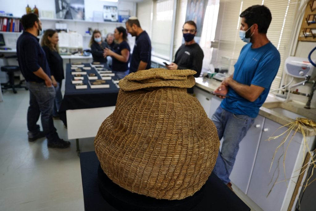 Israel Antiquities Authority archaeologists stand next to a 10,500-year-old basket dating back to the Neolithic period that was unearthed in Murabaat Cave in the Judean Desert, and is displayed at the IAA's Dead Sea conservation laboratory in Jerusalem. Photo by Menahem Kahana/AFP via Getty Images.