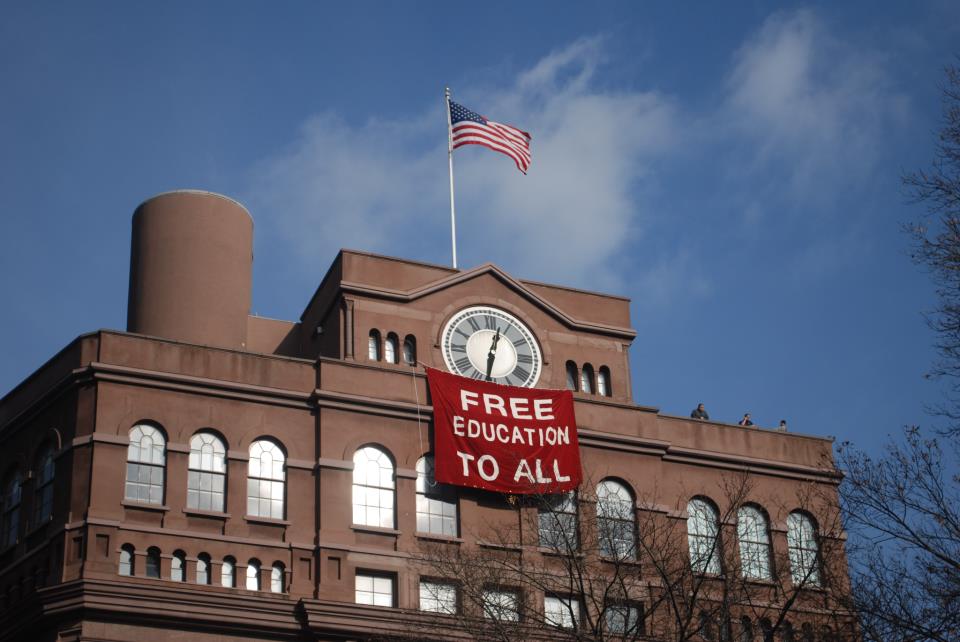 Students hang banner below the historic clock tower at Cooper Union in New York City during a 2012 occupation protesting implementing tuition in the historically free school. Photo by Free Cooper Union, Creative Commons Attribution-Share Alike 3.0 Unported license.