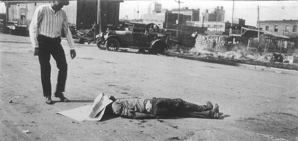 The body of an unidentified black victim of the Tulsa Race Massacre lies in the street as a white man stands over him, Tulsa, Oklahoma, June 1, 1921. Photo by Greenwood Cultural Center/Getty Images.