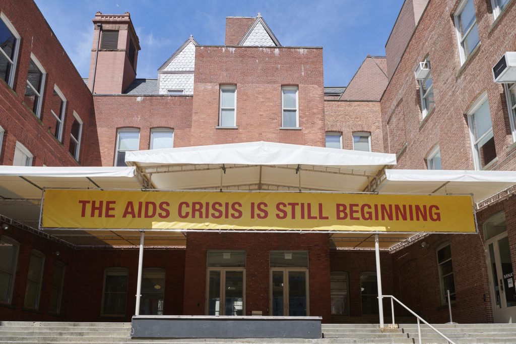 The banner leading up to MoMA PS1. Around 36 million people worldwide have the HIV virus. Only about half of them have access to life-saving drugs. Gregg Bordowitz, The AIDS Crisis is Still Beginning (2021). Image courtesy MoMA PS1. Photo: Kyle Knodell.