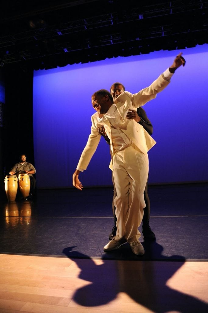 Birthing Dying Becoming Crip Wisdom (2016). Photo taken during the 2016 performance, Sins Invalid. Image description: On a dramatically lit stage, a Black man in a white suit stands front and center, his knees bent and his arms open. Right behind him, another Black man in a black suit holds him under his arms. In the background, a Black man sits at a pair of drums.
