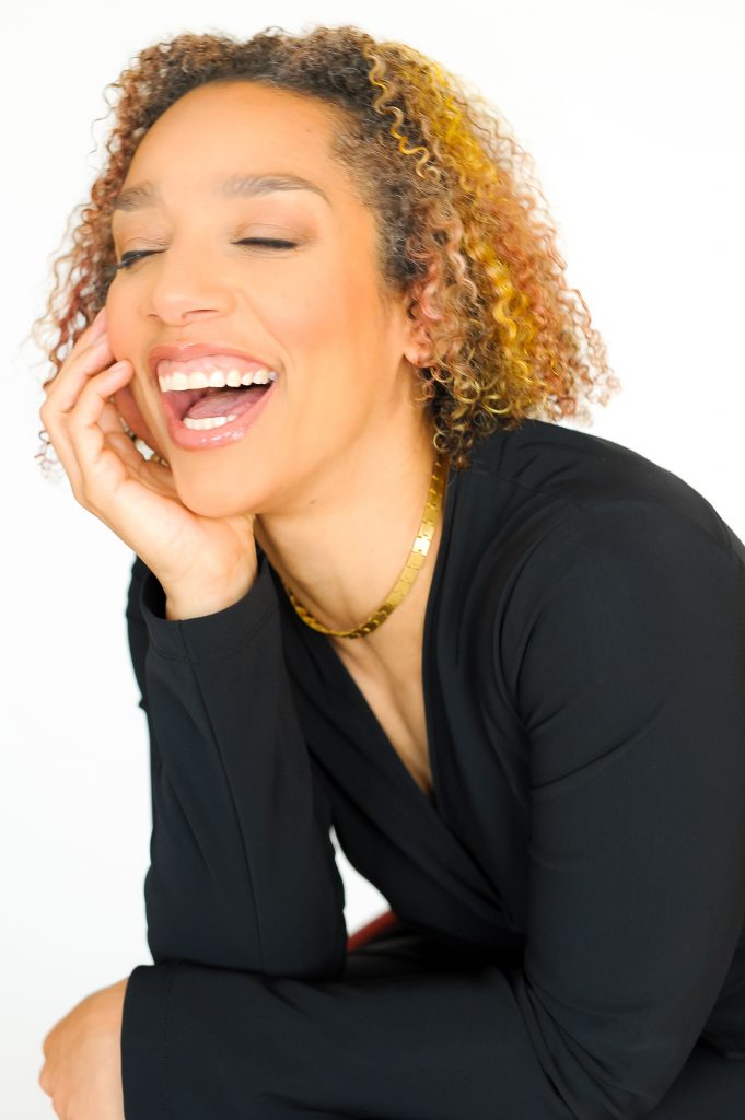 Alice Sheppard. Image description: A Black woman leans forward and smiles brightly, teeth showing and eyes closed, as she rests her chin in her palm. She has light brown skin, curly shoulder-length hair with subtle highlights, and wears a black blouse and sleek gold necklace.
