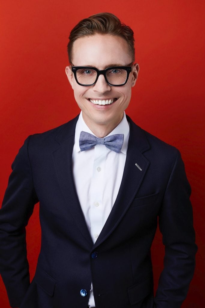 Lane Harwell. Image description: A nondisabled presenting white person with short blond hair and blue eyes behind black rim glasses smiles at the camera. They are wearing a white collared shirt with a light blue bowtie and a dark blue blazer against a red background.