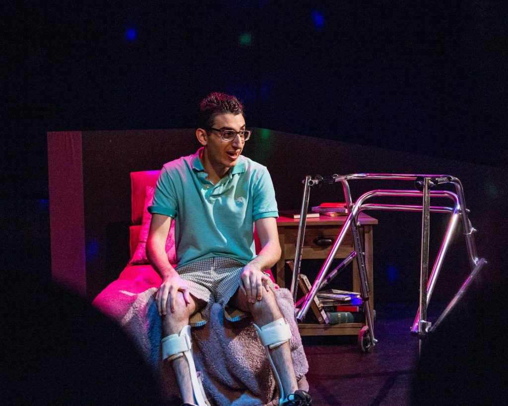 <i>Hi, Are You Single?</i> (2017). Production still from Ryan Haddad's solo play Hi, Are You Single? Photo by Michael Bernstein. Image description: Under cool stage lighting, Ryan Haddad sits at the end of a bed beside his metallic walker. He wears square glasses, a teal polo, patterned shorts, and lower leg braces. 