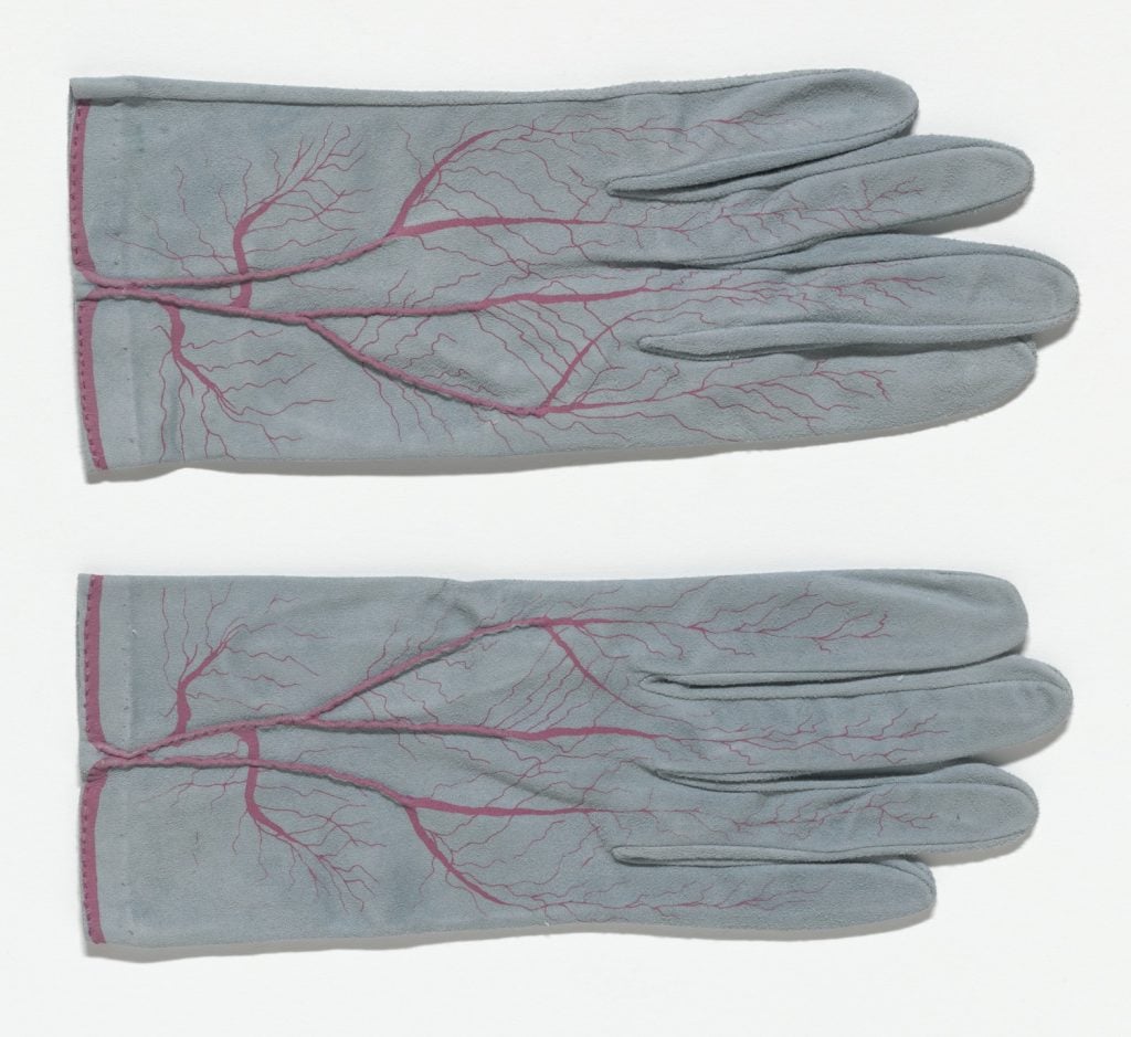 Meret Oppenheim, <i> Glove (for Parkett no. 4)</i> (1985). Courtesy of the Museum of Modern Art. ” srcset=”https://news.artnet.com/app/news-upload/2021/08/cri_000000266847-1024×939.jpg 1024w, https://news.artnet.com/app/news-upload/2021/08/cri_000000266847-300×275.jpg 300w, https://news.artnet.com/app/news-upload/2021/08/cri_000000266847-50×46.jpg 50w, https://news.artnet.com/app/news-upload/2021/08/cri_000000266847.jpg 1570w” width=”1024″ height=”939″></p>



<p>Meret Oppenheim, <em>Glove (for Parkett no. 4) </em>(1985). Courtesy of the Museum of Modern Art.</p>



<p>Oppenheim’s fascination with the body is also apparent in works like <em>Bees Knees—</em>a sterling silver platter with a pair of white women’s pumps bound together at the heels, which end in paper frills, like a bondage-inspired Norman Rockwell turkey dish. Oppenheim was also fascinated by hands and gloves, and many of her <em>objets d’art</em> used the garment in some way, with hand-painted and embroidered veins splayed on top of a pair of white leather gloves or a pair of hands ensconced in hirsute coverings from which red-painted nails poke out, like the extremities of a female werewolf in <em>Pelzhandschuhe</em>, 1936.</p>



<p>The artist was also a muse and model for her peers, most notably in <em>Erotique Voilée</em> a photographic series by Man Ray that placed a nude Oppenheim next to a printing press with one arm and hand covered in sticky dark paint. The lithe nude woman juxtaposed next to the large wheel with its iron spokes and phallic wooden handle creates a tableaux ripe for analysis.</p>



<p>Below, see some of Oppenheim’s most famous works.<img src=