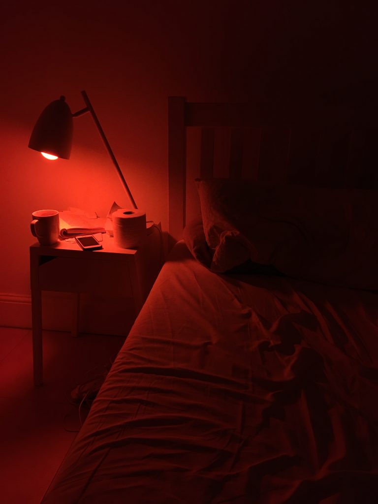 A dark bedroom lit by a glowing orange lamp on a bedside table. One side of the bed is visible, the sheets rumpled, the pillows smooshed, and the duvet pulled back. The bedside table also holds a cup of tea, an open book, a phone, and a roll of toilet tissue.
