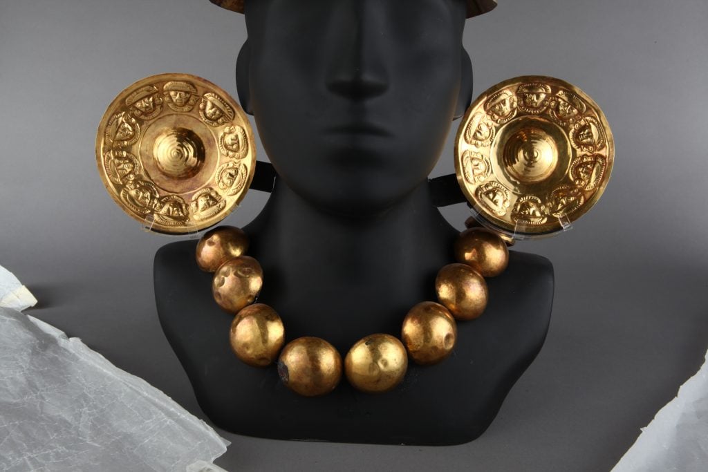 Gold-silver-copper alloy ear ornament with depiction of ten human heads with half-moon headdress, circular ear ornaments, and breastplate. Necklace of spherical beads made of a gold-silver-copper alloy. (ca. 1300 AD–1532 AD) Collection of the Museo Larco, Lima, Peru. Photo courtesy of World Heritage Exhibitions.