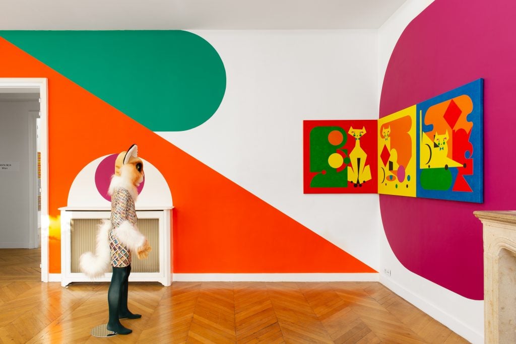 a fox-headed figure stands in the middle of a room against a backdrop of an orange, green, and fushcia mural