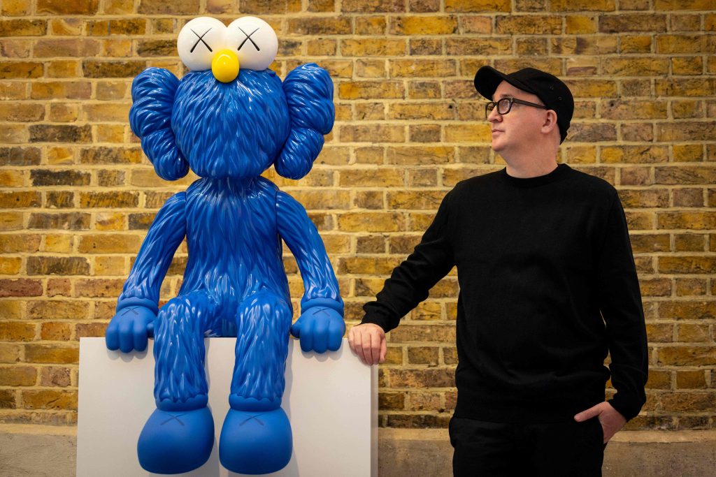 American artist KAWS, real name Brian Donnelly, poses with an artwork titled SEEING during a press preview for the exhibition 'KAWS: NEW FICTION' at the Serpentine North gallery in London on January 18, 2022. - RESTRICTED TO EDITORIAL USE - MANDATORY MENTION OF THE ARTIST UPON PUBLICATION - TO ILLUSTRATE THE EVENT AS SPECIFIED IN THE CAPTION (Photo by Tolga Akmen / AFP) / RESTRICTED TO EDITORIAL USE - MANDATORY MENTION OF THE ARTIST UPON PUBLICATION - TO ILLUSTRATE THE EVENT AS SPECIFIED IN THE CAPTION / RESTRICTED TO EDITORIAL USE - MANDATORY MENTION OF THE ARTIST UPON PUBLICATION - TO ILLUSTRATE THE EVENT AS SPECIFIED IN THE CAPTION (Photo by TOLGA AKMEN/AFP via Getty Images)